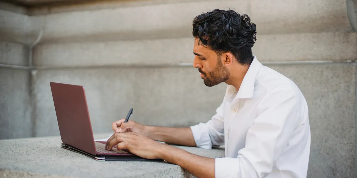 A man in a white shirt working at a small laptop on a grey concrete ledge, writing in a notebook beside the keyboard