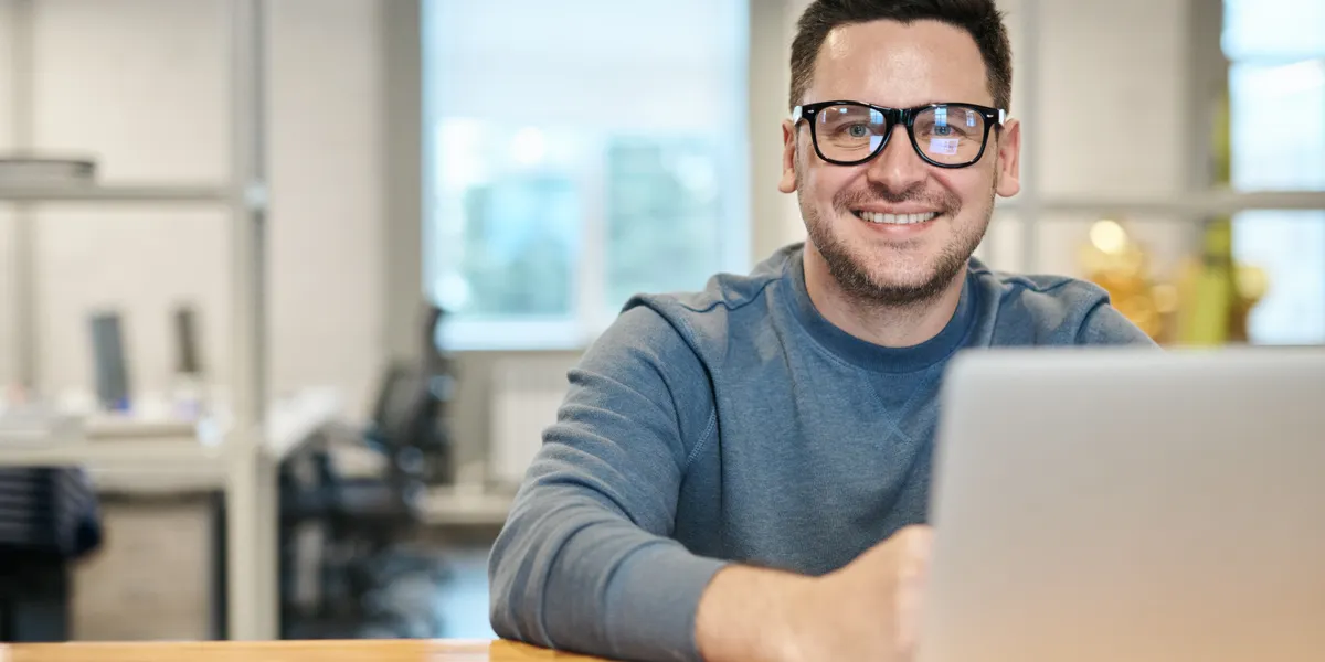 A bearded man in glasses and a blue-grey sweatshirt sitting at a wooden desk with an open laptop in a daylit open-plan office